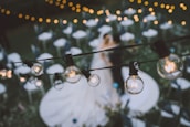 A softly focused wedding scene with hanging string lights in the foreground. The couple, blurred in the background, stands on a decorated area surrounded by chairs and greenery. The atmosphere suggests an outdoor evening event.