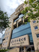 A tall, modern library building with a beige and blue color scheme, featuring large windows and architectural details like columns. The entrance has signage in both English and Chinese, with greenery in the foreground.