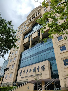 A tall, modern library building with a beige and blue color scheme, featuring large windows and architectural details like columns. The entrance has signage in both English and Chinese, with greenery in the foreground.