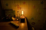 A dimly lit room with a wooden table displaying various objects, including a vintage style light bulb on a wooden base, a small toy car, a remote control, and some figurines. The background features a wallpaper with cartoonish animal and floral designs.