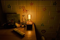 A dimly lit room with a wooden table displaying various objects, including a vintage style light bulb on a wooden base, a small toy car, a remote control, and some figurines. The background features a wallpaper with cartoonish animal and floral designs.