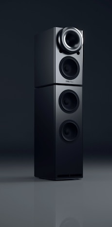 A sleek black audio speaker with a modern, minimalist design. The speaker consists of two stacked rectangular units, each featuring circular sound outputs. The top unit has a prominent cone-like speaker, while the bottom unit includes two additional circular drivers. The surface has a glossy finish, reflecting light subtly.