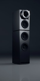 A sleek black audio speaker with a modern, minimalist design. The speaker consists of two stacked rectangular units, each featuring circular sound outputs. The top unit has a prominent cone-like speaker, while the bottom unit includes two additional circular drivers. The surface has a glossy finish, reflecting light subtly.