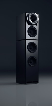 A sleek black audio speaker with a modern, minimalist design. The speaker consists of two stacked rectangular units, each featuring circular sound outputs. The top unit has a prominent cone-like speaker, while the bottom unit includes two additional circular drivers. The surface has a glossy finish, reflecting light subtly.