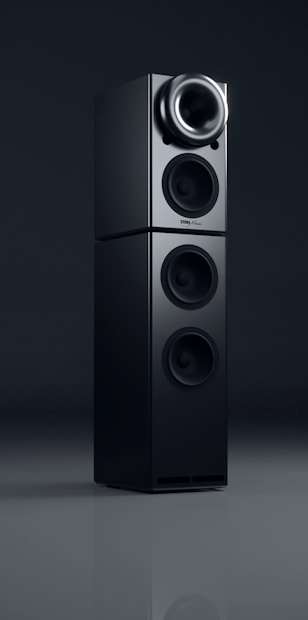 A sleek black audio speaker with a modern, minimalist design. The speaker consists of two stacked rectangular units, each featuring circular sound outputs. The top unit has a prominent cone-like speaker, while the bottom unit includes two additional circular drivers. The surface has a glossy finish, reflecting light subtly.