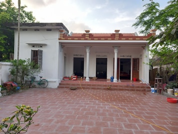 A traditional one-story house with a white exterior and a red tile roof. The house features three open doorways, each with decorative lintels. In front of the house is a tiled patio with a chicken walking across it. Various household objects, such as a bicycle, pots, and buckets, are scattered around. Lush greenery surrounds the area, including a palm tree and several bushes.