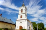 A tall, white church with a steeple topped by a cross stands prominently against a vibrant blue sky with scattered, wispy clouds. The church has arched windows with wooden shutters and a decorative entrance. Next to the church, a stone crucifix statue is visible surrounded by green foliage. A building with a grey tiled roof is adjacent on the left.
