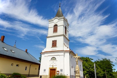 A tall, white church with a steeple topped by a cross stands prominently against a vibrant blue sky with scattered, wispy clouds. The church has arched windows with wooden shutters and a decorative entrance. Next to the church, a stone crucifix statue is visible surrounded by green foliage. A building with a grey tiled roof is adjacent on the left.