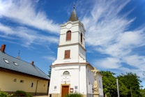 A tall, white church with a steeple topped by a cross stands prominently against a vibrant blue sky with scattered, wispy clouds. The church has arched windows with wooden shutters and a decorative entrance. Next to the church, a stone crucifix statue is visible surrounded by green foliage. A building with a grey tiled roof is adjacent on the left.