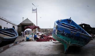 Several blue and turquoise fishing boats are docked on a concrete pier, with people nearby working on fishing nets. The scene includes an old stone building with a pointy roof and a signpost above. Seagulls are flying in the overcast sky, indicating a coastal environment.
