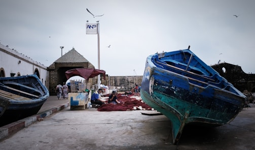 Several blue and turquoise fishing boats are docked on a concrete pier, with people nearby working on fishing nets. The scene includes an old stone building with a pointy roof and a signpost above. Seagulls are flying in the overcast sky, indicating a coastal environment.