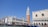 A sunny view of the famous Campanile and Doge's Palace in Venice, Italy, with gondolas lined up along the canal. The architecture features ornate columns, arched windows, and a bright tower with a green pointed roof. The sky is clear and blue, enhancing the vibrant atmosphere.