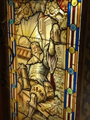 A stained glass window depicts a bearded king wearing a crown, holding up a mug of beer in one hand and a scepter in the other. The background features a castle and a sun with a face, surrounded by vines and flowers. The scene is framed with colorful geometric patterns.