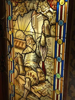 A stained glass window depicts a bearded king wearing a crown, holding up a mug of beer in one hand and a scepter in the other. The background features a castle and a sun with a face, surrounded by vines and flowers. The scene is framed with colorful geometric patterns.