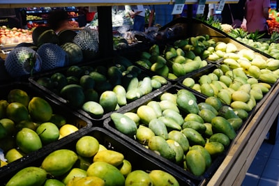 A supermarket display filled with a variety of fresh mangoes, arranged in black trays. The mangoes range in color from green to ripe yellow, with some other fruits visible in the background. Several people are partially visible, shopping in the well-lit produce section.