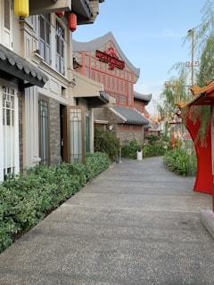 A pathway lined with green plants leads to a building with traditional Asian architectural elements, including a prominent red sign. Ornate details like hanging lanterns and decorative roof edges are visible, surrounded by lush greenery.