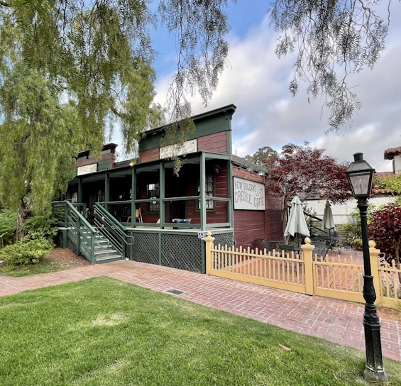 A quaint wooden building with green trim, and a sign reading 'Creole Café' is surrounded by greenery and a small outdoor seating area with umbrellas. A classic street lamp and a yellow picket fence enhance the charm. The scene includes lush grass in the foreground and trees in the background under a partly cloudy sky.