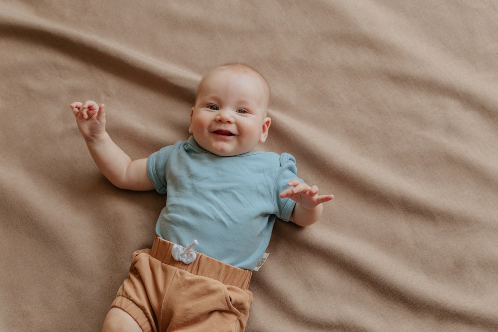bebê na camisa azul e calções marrons deitado sobre o tecido marrom