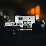 A vintage Yashica camera and a modern Canon camera are placed on a dark surface in front of a laptop displaying an Instagram gallery featuring various black and white images. Soft, warm light glows in the background, adding an atmospheric touch.