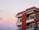 A modern apartment building with a red-tiled roof and balconies. The sky is a gradient of soft pastel colors, transitioning from light pink to purple. The building features brick and concrete materials with glass windows and metal railings.