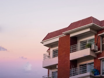 A modern apartment building with a red-tiled roof and balconies. The sky is a gradient of soft pastel colors, transitioning from light pink to purple. The building features brick and concrete materials with glass windows and metal railings.
