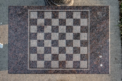 A granite chessboard inlaid on a stone surface. The board displays a classic grid pattern with alternating darker and lighter squares. It sits on a paved surface, surrounded by natural elements like grass. The texture of the board is rough, suggesting outdoor use.