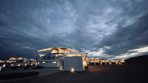 A series of modern, white buildings are illuminated under a dramatic and cloudy sky, with soft lighting coming from the structures. The architecture features angular, futuristic designs.