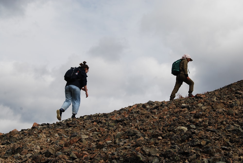 homme en veste noire et jean bleu debout sur une colline rocheuse sous un ciel nuageux blanc