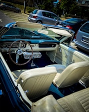A vintage convertible car with a classic steering wheel and dashboard is parked in an outdoor lot. The interior features light-colored leather seats. Several modern cars are parked nearby, and trees with a building backdrop can be seen in the background.