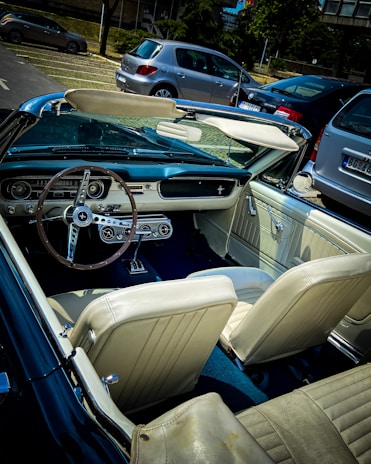 A vintage convertible car with a classic steering wheel and dashboard is parked in an outdoor lot. The interior features light-colored leather seats. Several modern cars are parked nearby, and trees with a building backdrop can be seen in the background.