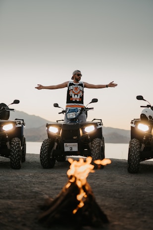 A person stands confidently on an ATV with arms spread wide, surrounded by two other ATVs. In the foreground, a small campfire emits a warm glow. The setting appears to be outdoors, with mountains and a body of water in the background during sunset.
