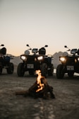 A group of all-terrain vehicles (ATVs) parked on a dirt surface with a small campfire in front. The background features a mountainous landscape under a twilight sky, adding a sense of adventure to the scene. The headlights of the ATVs are on, casting light towards the viewer.