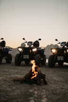 A group of all-terrain vehicles (ATVs) parked on a dirt surface with a small campfire in front. The background features a mountainous landscape under a twilight sky, adding a sense of adventure to the scene. The headlights of the ATVs are on, casting light towards the viewer.