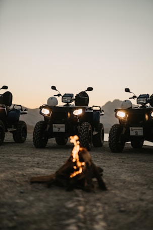 A group of all-terrain vehicles (ATVs) parked on a dirt surface with a small campfire in front. The background features a mountainous landscape under a twilight sky, adding a sense of adventure to the scene. The headlights of the ATVs are on, casting light towards the viewer.