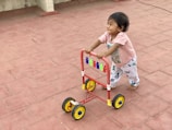 A young child is joyfully pushing a colorful walker on a red-tiled surface. The child is bare-footed, wearing a light pink shirt and patterned pants. The walker is bright with red, yellow, and blue accents, creating a playful atmosphere.