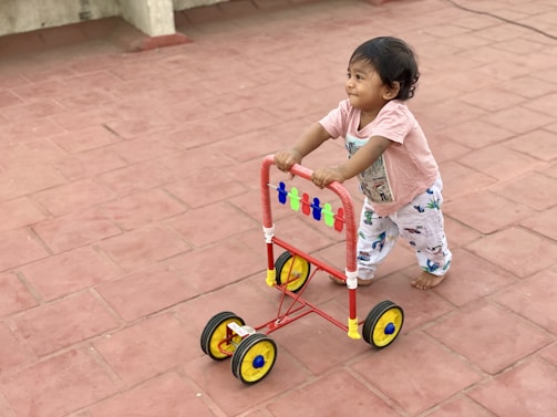 A young child is joyfully pushing a colorful walker on a red-tiled surface. The child is bare-footed, wearing a light pink shirt and patterned pants. The walker is bright with red, yellow, and blue accents, creating a playful atmosphere.