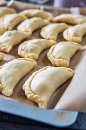 A baking sheet lined with parchment paper holds multiple raw empanadas, neatly arranged in rows. The empanadas have a crimped edge, indicating they are ready to be baked, and the light reflects softly off their surface.