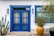 A bright blue wooden door is framed by two wall-mounted lanterns. To the left, there is a tall, green cactus with multiple arms and small flowers. On the right, a large orange-yellow ceramic pot with purple flowering plants is placed in front of a window with matching blue trim. The house exterior is painted white, creating a vibrant contrast with the door and window frames.