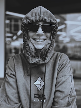 A person wearing a hoodie is smiling while looking directly at the camera. They are also wearing large dark sunglasses. The hoodie has a design featuring a stylized image and some text. The background appears to be an indoor setting with blurred lights.