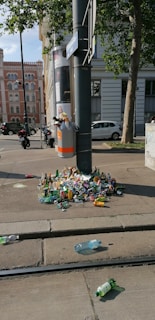 A street scene with a pole on the sidewalk, which has become a makeshift trash collection point. Trash, including plastic bottles and cans, is scattered around and overflowing from the bin. The area appears to be in an urban setting with a backdrop of buildings and some parked vehicles. Trees and a clear sky suggest daylight.
