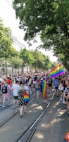 A large crowd of people gathered in a tree-lined street, many holding or wearing rainbow flags and accessories. The atmosphere appears festive and celebratory, with diverse participants engaged in a parade or march. Buildings line the street in the background.