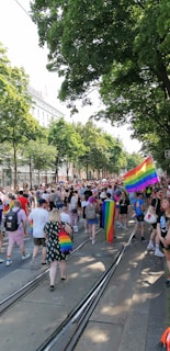 A large crowd of people gathered in a tree-lined street, many holding or wearing rainbow flags and accessories. The atmosphere appears festive and celebratory, with diverse participants engaged in a parade or march. Buildings line the street in the background.