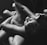 A grayscale image depicting an intimate and closely cropped view of two people entwined together. The focus is on their intertwined limbs and the serene expression on the visible face. The lighting creates deep contrasts and highlights the contours of their bodies.