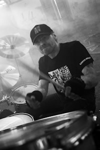 A musician, wearing a cap and a graphic t-shirt, is energetically playing a drum set in a slightly smoky, dimly lit environment. The cymbals and drumsticks are prominently visible, emphasizing the focus on music.