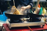 A person is preparing noodles in a wok on a red table with a stove beneath. Steam rises from the noodles as they are being stirred using a metal tool. Another wok is placed beside the one in use, and there are blurred background elements suggesting a busy kitchen environment.