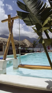 A tropical resort setting featuring a swing hanging over a swimming pool. The pool water is clear and turquoise, surrounded by thatched-roof structures. A large green leaf is prominently in the foreground, with mountains and clouds visible in the background. There are lounge chairs and people relaxing by the poolside.
