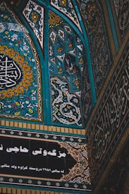Intricate tilework featuring elaborate geometric patterns and floral motifs, dominated by shades of blue, yellow, and white. Arabic or Persian script adorns sections of the design, suggesting cultural or religious significance.