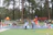 Parents watching happily as their kids play on a large green and beige inflatable slide in a forest setting.