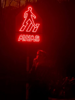 A glowing red warning sign with the word 'AWAS' and an outline of a person descending steps. Below the sign, a person is partially visible, standing in the dark and holding something near their face.