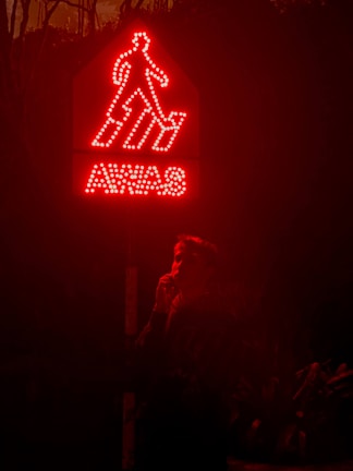 A glowing red warning sign with the word 'AWAS' and an outline of a person descending steps. Below the sign, a person is partially visible, standing in the dark and holding something near their face.